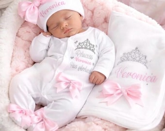 Peut inclure: Un ensemble de grenouillère et bonnet pour bébé blanc avec des nœuds roses et des motifs de diadème à paillettes argentées. Le texte indique Princess Veronica Has Arrived sur la grenouillère et Veronica sur le bonnet et une couverture.
