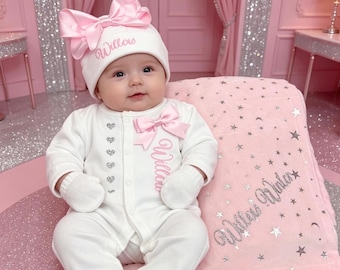 May include: A baby in a white outfit with pink bows and the name "Willow." The outfit includes a hat, onesie, and mittens. The baby sits on a pink blanket with stars and the name "Willow Winter." The background is a pink room with silver accents.