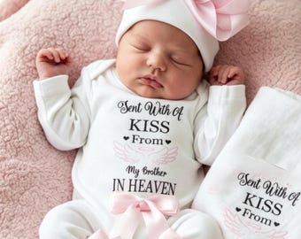 May include: A white long-sleeved baby bodysuit and matching hat set featuring a large pink satin bow on the hat and smaller pink bows on the legs. The text reads Sent With A KISS From My Brother IN HEAVEN with pink angel wings and hearts.