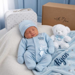 Peut inclure: Une grenouillère bleu clair pour bébé avec un bonnet assorti, portant le texte brodé "Hello World I'm Ryan" et une couronne. Un ours en peluche blanc avec un nœud bleu et le nom "Ryan" est posé près d'une couverture bleue brodée "Ryan".