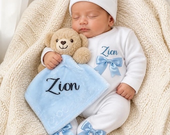 May include: A white baby onesie and matching beanie set featuring the name Zion embroidered in blue, accented with light blue satin bows on the chest and feet, paired with a light blue plush security blanket embroidered with the name Zion.