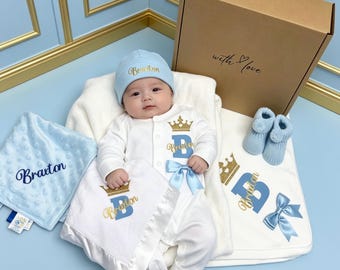 May include: A baby gift set featuring a light blue beanie, a white long-sleeve onesie with a gold crown and blue letter B, a light blue textured security blanket, a white blanket with satin trim, and blue booties, all personalised with the name Braxton.