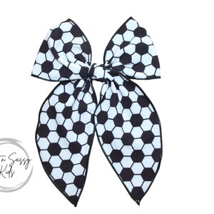 May include: A large hair bow featuring a soccer ball pattern in black and white. The bow is tied in the center and has long, flowing tails. The bow is likely for a child and is set against a white background.
