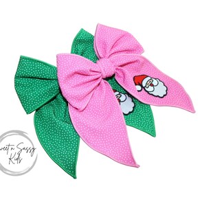 May include: Three fabric hair bows. One pink bow with white polka dots and a Santa Claus graphic, and two green bows with white polka dots. The bows are arranged on a white background. The bows are for children.