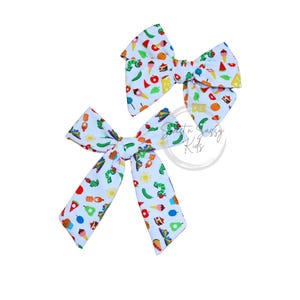 May include: Two white hair bows featuring a colorful pattern of small illustrations including green caterpillars, red apples, yellow suns, ice cream cones, and various food items. The bows have a knotted center and a fabric texture with a repeating print.