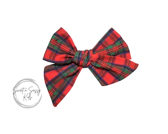 May include: A red, green, and navy plaid bow. The bow is tied in the center and has two large loops on either side. The bow is a hair accessory.