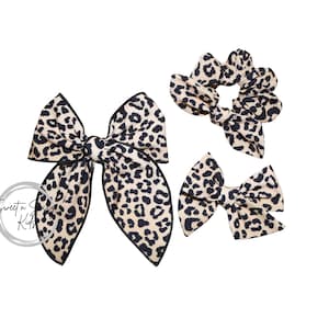 May include: A set of three hair accessories: a large bow, a small bow, and a scrunchie. All items feature a tan and black leopard print pattern with black trim. The accessories are designed for hair styling.