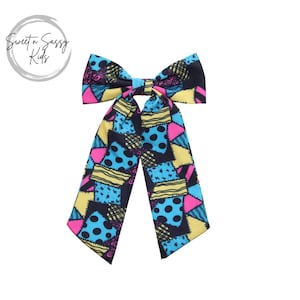 May include: A large, colorful bow with a patchwork design in shades of blue, yellow, pink, and black. The bow is tied in a knot and has two long tails.