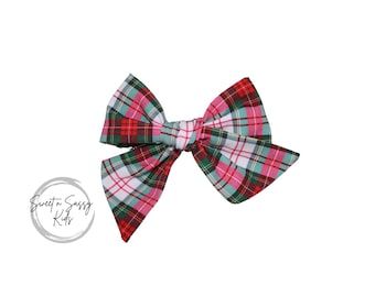 May include: A large, handmade hair bow with a classic plaid pattern in red, green, pink, and white. The bow is tied in the center and has two large loops on each side. The bow is a festive accessory.