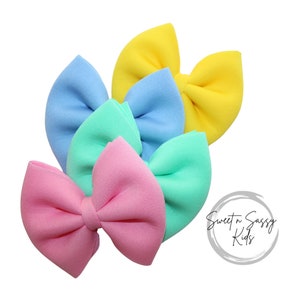 May include: Four pastel colored fabric hair bows. The bows are in shades of pink, blue, green, and yellow. The bows are arranged in a stack, with the pink bow on the bottom and the yellow bow on the top. The bows are all tied in a bow shape. The bows are all made of a soft, velvety fabric. The bows are all about 3 inches in diameter. Sweet n Sassy Kids is written in a circle on the right side of the image.