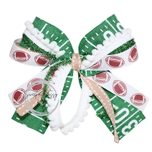 May include: A decorative hair bow featuring a football theme. The bow has green and white ribbons with a football field design and football graphics. White pom-poms and a light brown accent ribbon add detail. The text "Sweet & Sassy" is visible.