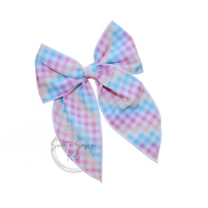 May include: A large, pastel-colored hair bow with a gingham pattern. The bow features shades of pink, yellow, blue, and purple. The bow is tied in the center and has long, flowing tails. The text "Sweet n Sassy Kids" is visible.