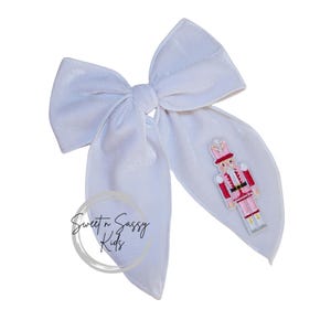May include: A large, white velvet hair bow with a decorative embroidered pink and red Nutcracker soldier. The bow features long, flowing tails and a central knot. The text "Sweet n Sassy Kids" is printed on the bow.