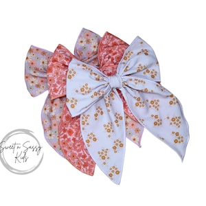 May include: Four fabric hair bows in shades of pink and white, each with a floral pattern. The bows are arranged in a layered display against a white background. The bows have a soft, feminine aesthetic.