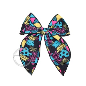 May include: A large fabric hair bow featuring a patchwork pattern with black, bright blue, yellow, and hot pink sections. The design includes various geometric shapes, polka dots, and swirling line patterns across the entire surface of the bow.