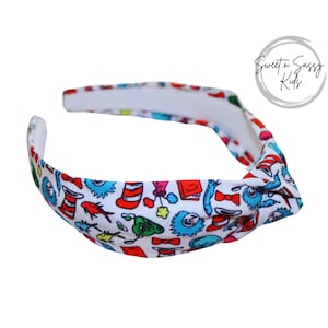May include: A white headband with a knotted design. The headband is covered in a colorful print featuring characters and objects from the book "The Cat in the Hat".