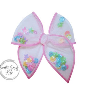May include: A white tulle bow with a pink trim and colorful sequins. The bow is decorated with pastel-colored sequins in various shapes, including flowers and circles.