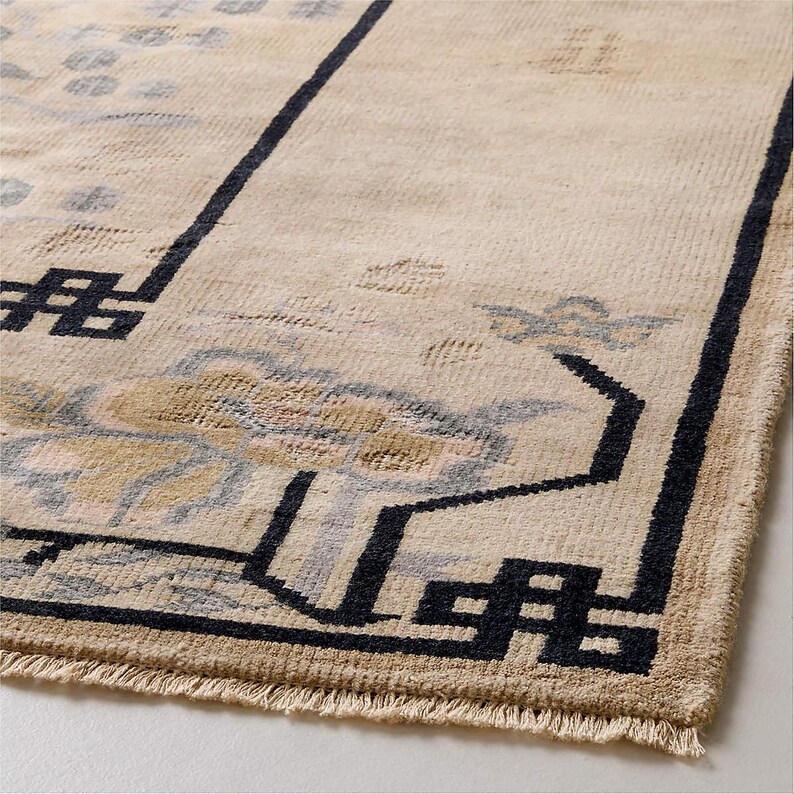 May include: Beige area rug with a geometric design. The rug features a central floral pattern in shades of beige, blue, and gold, framed by a black border with geometric shapes. The rug has a fringe along one edge.