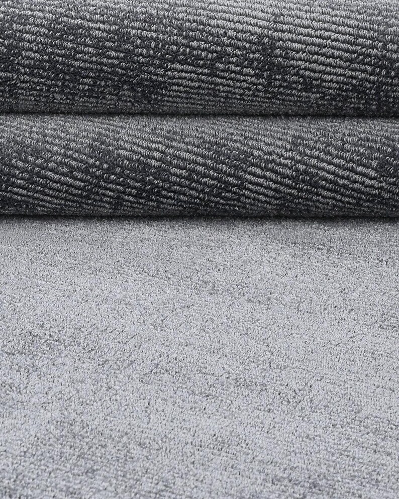 May include: Close-up of a gray carpet with a textured surface. The carpet is rolled up in two layers, revealing the woven pattern. The bottom layer is a flat, light gray surface.