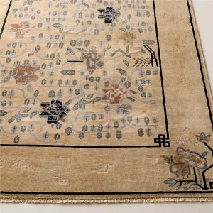 May include: Beige area rug with a floral pattern in shades of brown, gray, and black. The rug has a black border and a fringed edge. The design features stylized flowers and leaves.