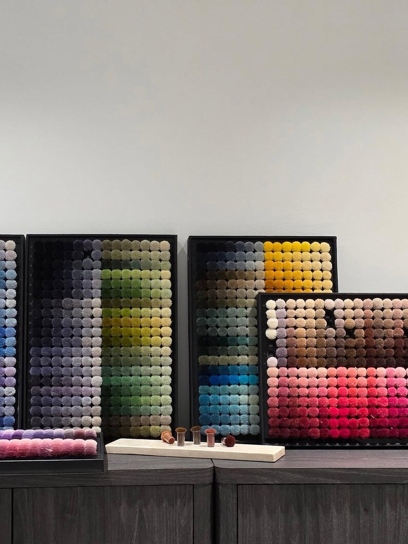 May include: A display of framed pom-pom art in various color gradients. The pom-poms are arranged in a grid pattern, showcasing a range of hues from black to yellow and pink to red. Several small, round objects are on a wooden display.