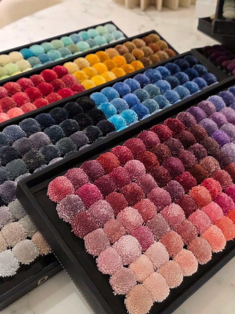May include: A collection of black rectangular trays filled with rows of small, round, fuzzy pom-poms in a wide spectrum of colors. The pom-poms are arranged in gradients, featuring shades of pink, red, purple, blue, yellow, orange, and gray.
