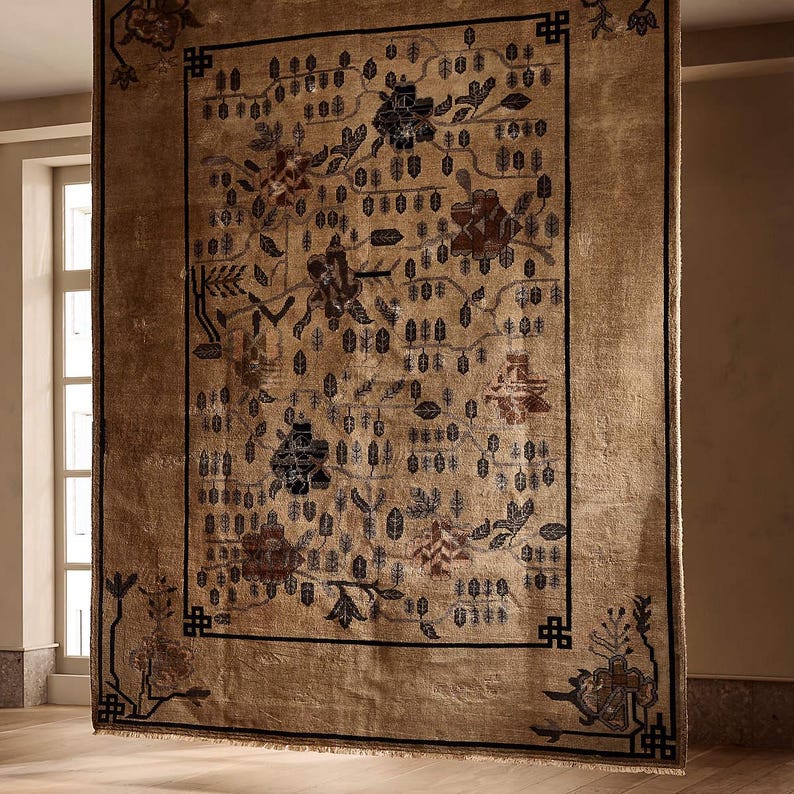 May include: A large, rectangular rug with a beige background and a detailed floral pattern in shades of brown, blue, and black. The rug has a black border and is hanging in front of a window. The rug's design includes flowers, leaves, and geometric shapes.