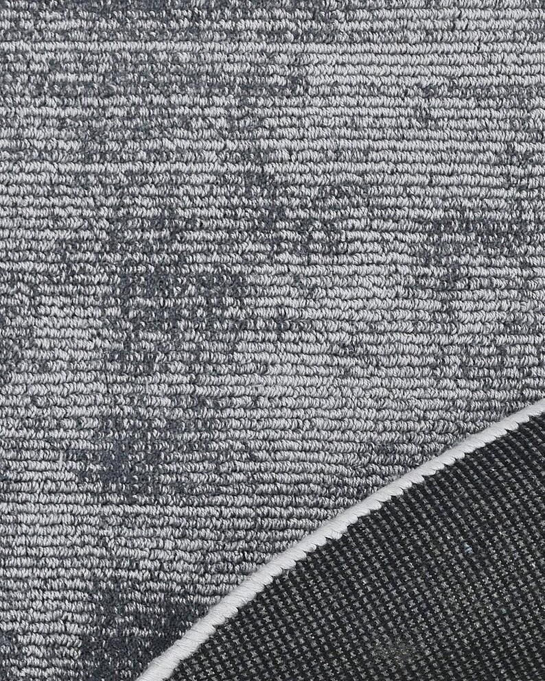 May include: Close-up of a gray rug with a textured surface. The rug has a pattern of darker gray spots and a white border. The rug's texture is visible, with a mix of light and dark gray fibers.