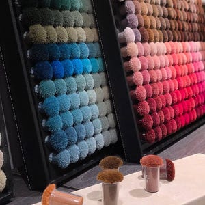 May include: A color sample display board featuring a grid of small, round, textured fabric tufts in various shades of blue, green, and gray, alongside a second board with warm tones of pink, red, and brown, with individual tuft samples in front.