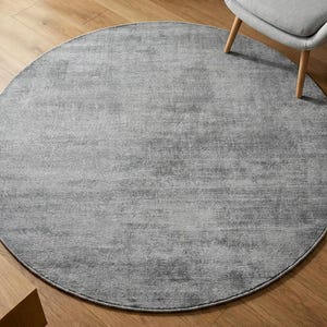 May include: A large, round, gray rug with a textured surface. The rug is placed on a hardwood floor, with a gray chair and a wooden side table visible in the background. The rug's design is simple and modern.