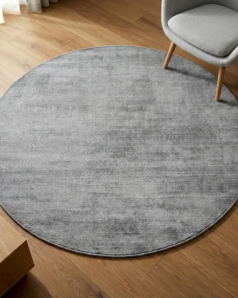 May include: A large, round, gray rug with a textured surface. The rug is placed on a hardwood floor, with a gray chair and a wooden side table visible in the background. The rug's design is simple and modern.