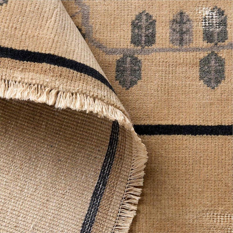 May include: Beige and black area rug with a geometric pattern. The rug has a rolled-up edge revealing a striped design and fringe. The pattern includes dark gray tree-like shapes and a black horizontal stripe.