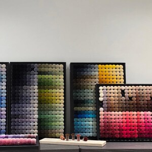 May include: A display of framed pom-pom color swatches in various shades, including black, gray, green, blue, yellow, and pink. The pom-poms are arranged in a grid pattern, showcasing a range of hues. Several small makeup brushes are on a wooden board.