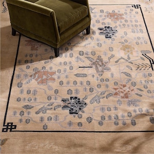 May include: Beige area rug with a floral pattern in shades of orange, gray, and black. The rug has a black border and is partially covered by a green armchair. The rug's design includes stylized flowers and leaf motifs.