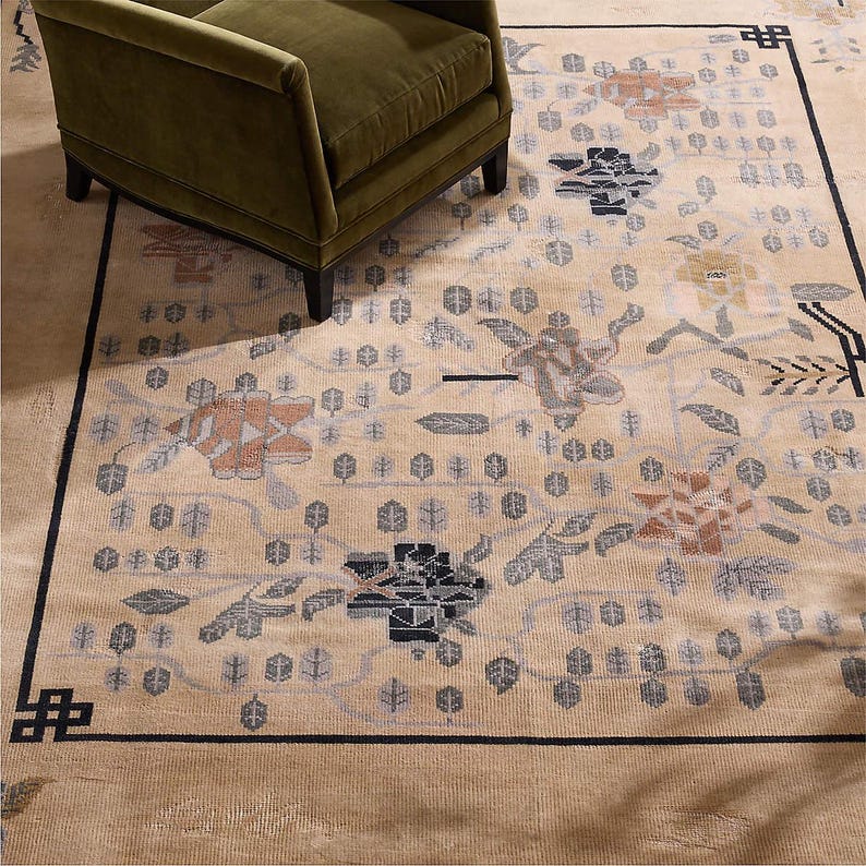 May include: Beige area rug with a floral pattern in shades of orange, gray, and black. The rug has a black border and is partially covered by a green armchair. The rug's design includes stylized flowers and leaf motifs.