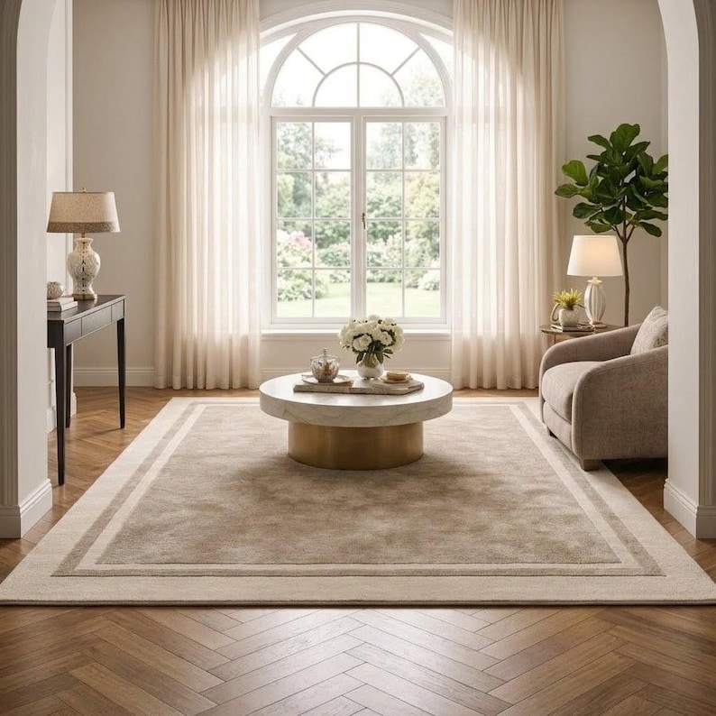 May include: A rectangular area rug featuring a solid beige center bordered by a lighter cream frame. The rug is positioned on a brown herringbone wood floor in a room with a round coffee table, a beige armchair, and two table lamps.