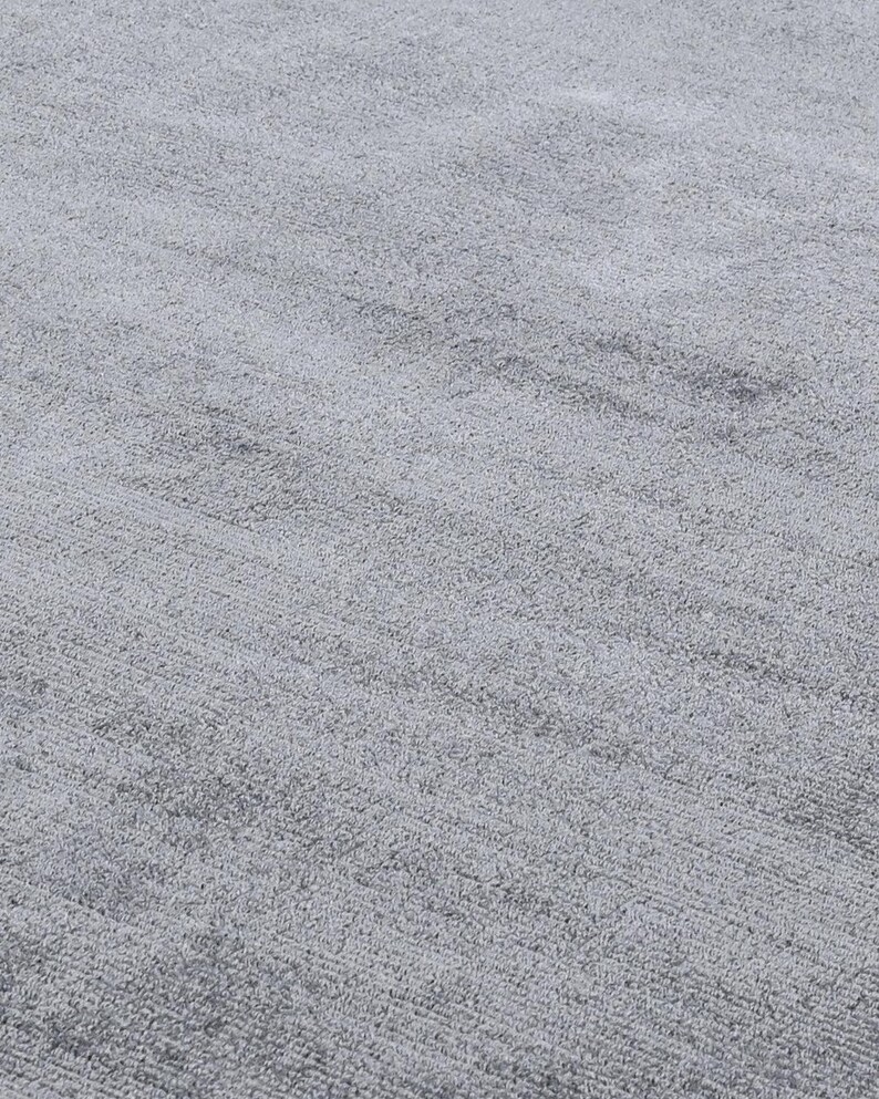 May include: Close-up of a light gray, textured rug. The rug has a soft, plush appearance with subtle variations in tone, creating a sense of depth and dimension. The fibers are densely packed, suggesting a comfortable and durable surface.