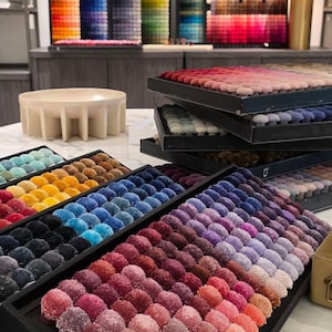 May include: A collection of rectangular sample trays containing rows of small, round, textured fabric tufts in a wide spectrum of colors, including shades of red, blue, purple, yellow, and neutral tones, arranged in organized color gradients.