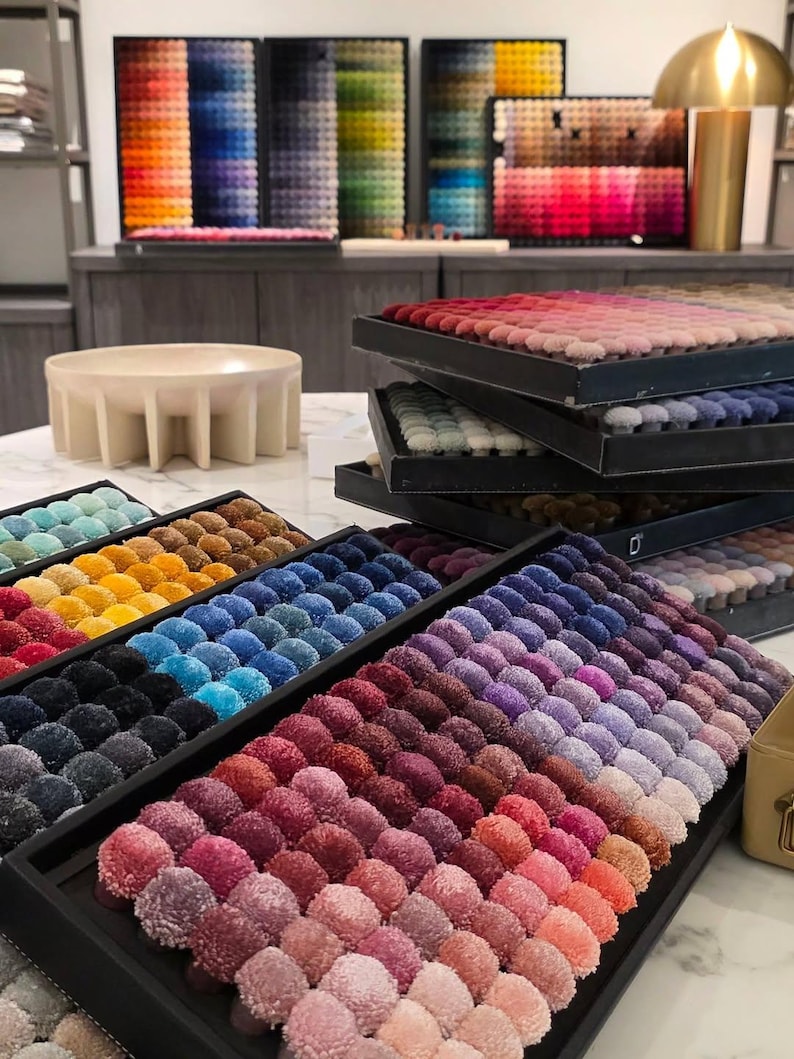 May include: A collection of rectangular sample trays containing rows of small, round, textured fabric tufts in a wide spectrum of colors, including shades of red, blue, purple, yellow, and neutral tones, arranged in organized color gradients.