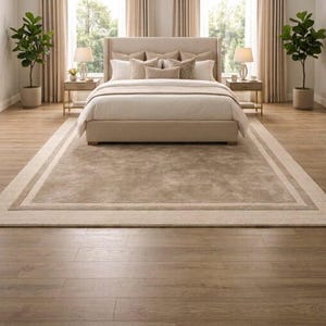 May include: A beige upholstered bed frame with a tufted headboard, dressed in white bedding and multiple decorative pillows. The bed is centered on a large, rectangular area rug with a border, flanked by two wooden nightstands with lamps.