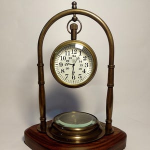 Puede incluir: Un reloj de escritorio de estilo vintage con esfera redonda y base de lupa. El reloj tiene una esfera blanca con números y manecillas negras, y un marco color latón. La base es de madera oscura.