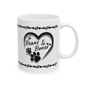 Può includere: Tazza in ceramica bianca con un design a cuore nero che include le parole "Beans & Bones" e impronte di zampe. La tazza ha un manico e un bordo di ossa e chicchi di caffè.