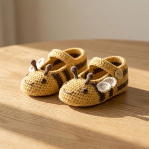 Puede incluir: Un par de zapatos de bebé de ganchillo hechos a mano con diseño de abeja. Los zapatos son amarillos con rayas marrones y presentan antenas, alas y cierre de botón. Están sobre una superficie de madera.