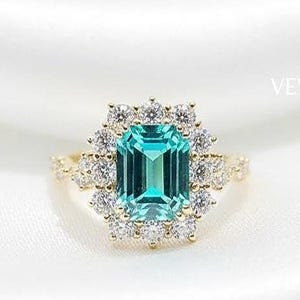 May include: A gold ring featuring a rectangular, teal-colored gemstone surrounded by a halo of smaller, clear stones. The ring is set against a white, silky background, with the brand name "VEVA JEWEL" visible.