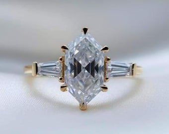 May include: A marquise-cut diamond engagement ring with a gold band. The ring features a large, clear marquise-cut center stone, flanked by two smaller baguette diamonds. The ring is set against a white, silky background.