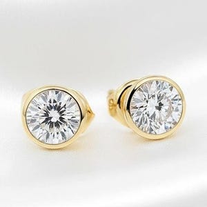 May include: A pair of gold stud earrings with a round, clear gemstone. The earrings are set in a bezel setting, and the gemstones sparkle against the white background. The earrings are a classic and elegant accessory.