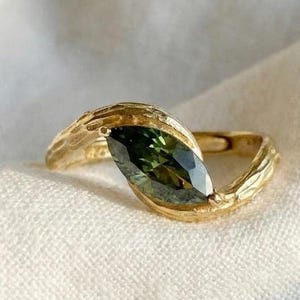 May include: A gold ring with a textured band and a dark green, oval-shaped gemstone. The ring's design features a flowing, organic shape, with the gemstone set in the center. The ring is displayed on a white, textured surface. The brand name "VEVA JEWEL" is visible.