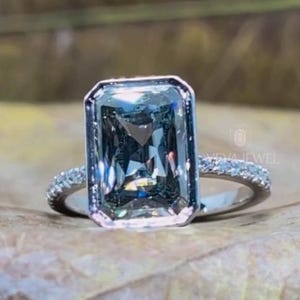May include: A silver ring with a large, rectangular, light blue gemstone. The band is encrusted with small, clear stones. The ring is set on a brown leaf, with the words "VEVA JEWEL" at the bottom.