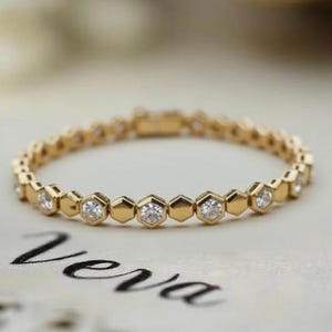 May include: A delicate gold bracelet with a repeating pattern of hexagonal links, each set with a small, sparkling diamond. The bracelet is displayed on a surface with the handwritten word "Vera".