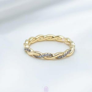 May include: A gold-colored ring with a braided design and small, clear stones. The ring is set against a white background, showcasing the intricate details of the jewelry. The ring is a piece of fashion jewelry.